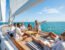 Diverse group of friends on a sailboat deck, laughing, playing a board game, and relaxing under a bright blue sky on turquoise waters.