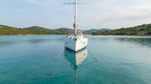 A white sailboat is perfectly centered in a clear, turquoise bay, securely held by two visible anchor lines. Green hills line the background under a bright sky.