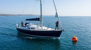 A sleek dark-hulled monohull sailboat with two crew members approaches a red and yellow mooring buoy on calm blue water under a clear sky.