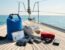 Équipement essentiel (sac étanche, licence, trousse de secours, chaussures) sur le pont d'un voilier en mer sous un ciel bleu.
