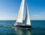 Un voilier moderne à coque sombre nommé "Aurora" naviguant à la voile sur une mer bleue sous un ciel dégagé.